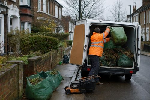 Worker lifting sacks of garden waste and cut branches into a van on a London residential street